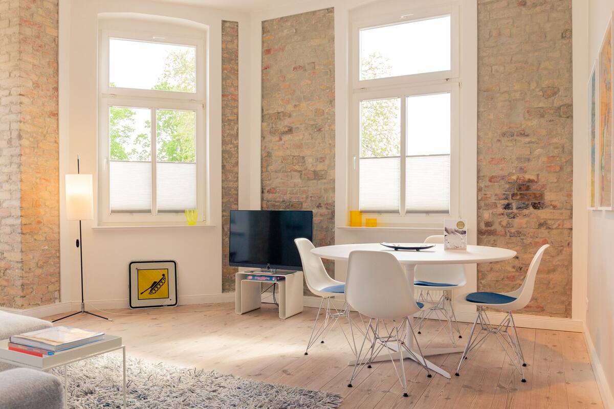 Een moderne woonkamer met een bakstenen muur, grote ramen, een flatscreen-televisie en een ronde eettafel met witte stoelen.