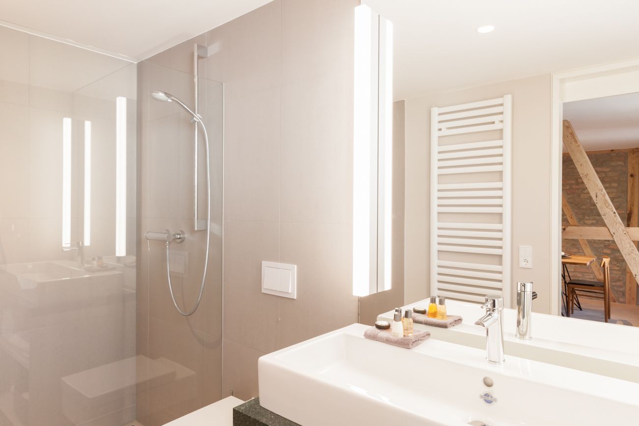 Een moderne badkamer met een douchecabine van glas, een groot wit wastafelmeubel met spiegel, en een houten trap op de achtergrond.