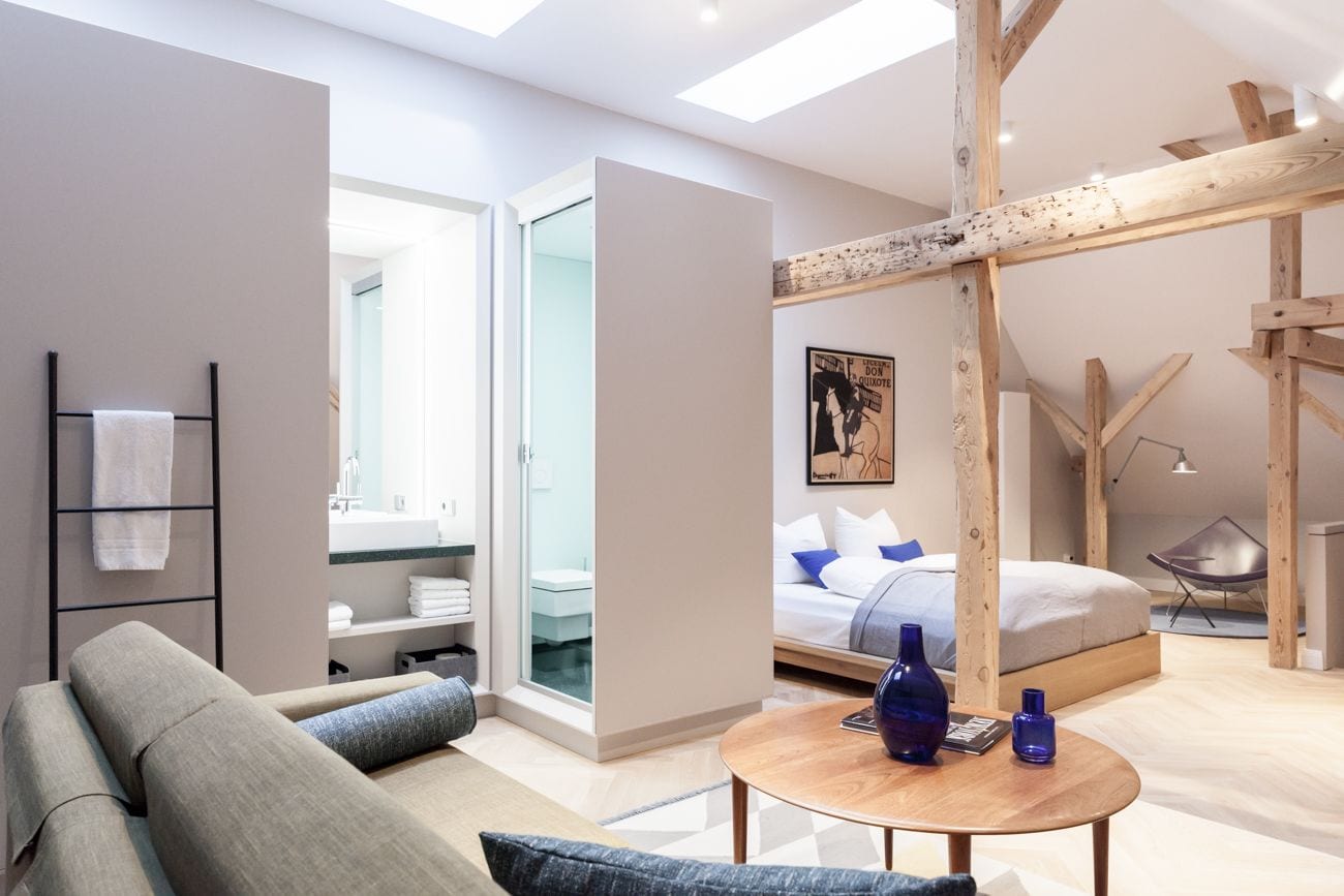 Habitación de hotel moderna con cama, techo de madera, una mesa redonda de madera y vista al baño.