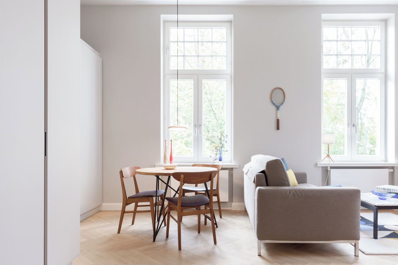 A bright, modern living room with a dining area, large windows, and a tennis racket on the wall.