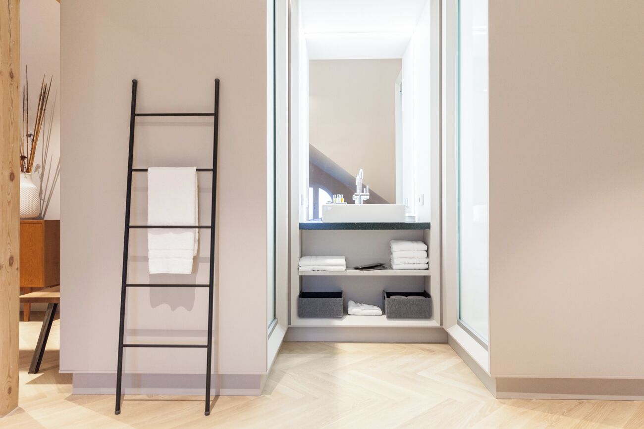 A modern bathroom with a wooden floor, a black metal ladder, and a built-in shelf with folded towels and storage boxes.