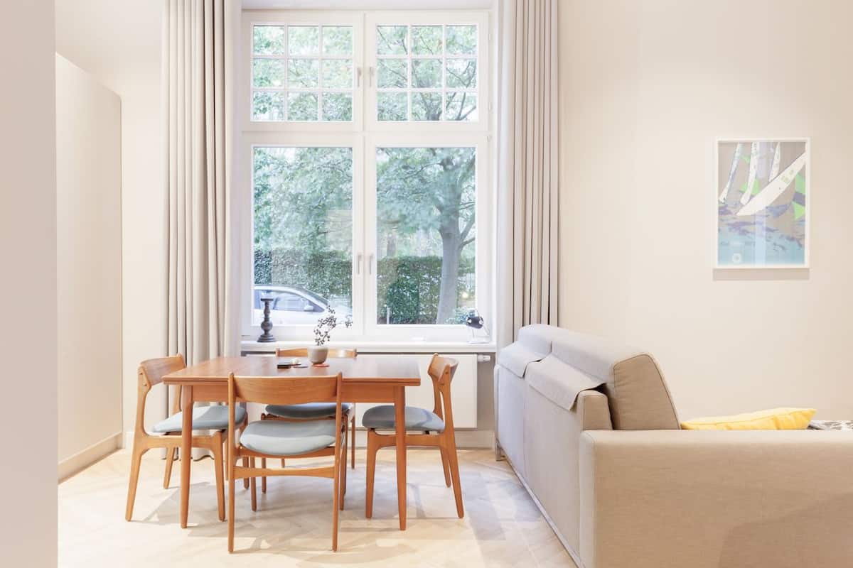 A modern living room with a dining table and chairs, a sofa, and a large window with curtains.