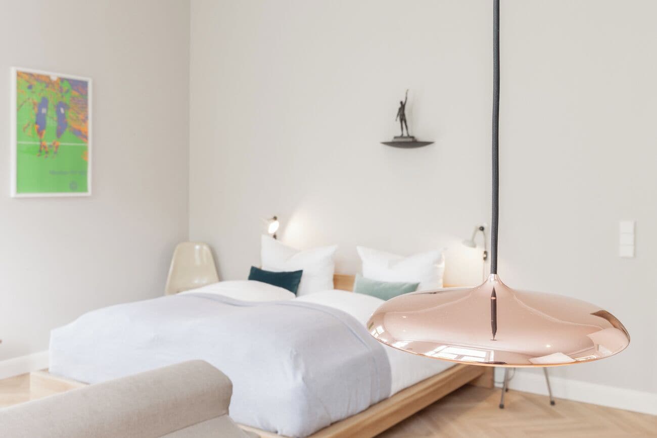 A modern bedroom with a bed, bedside lamps, a hanging light fixture, and a wall-mounted shelf.