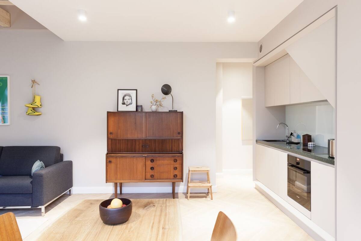 A modern living room with a dark grey sofa, a wooden cabinet, and a small table with a bowl of fruit. A kitchen with white cabinets and a countertop is visible to the right.