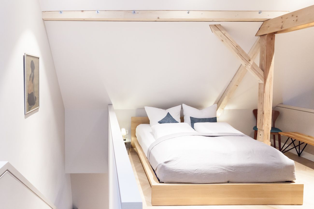 Un dormitorio con estructura de cama de madera, ropa de cama blanca y techo inclinado con vigas a la vista.