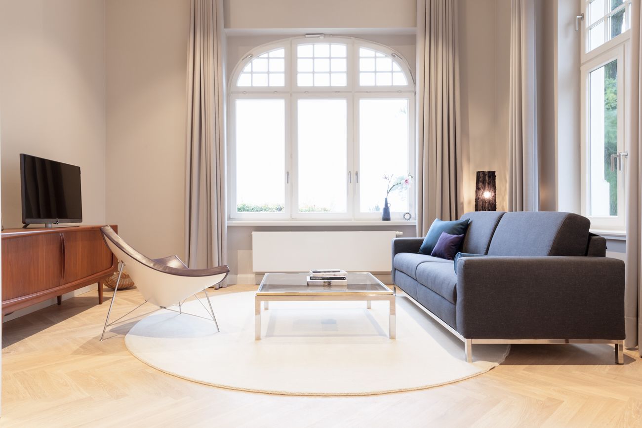 Ein modernes Wohnzimmer mit einer Couch, einem Glastisch, einem Fernseher auf einem Holzschrank und einem großen Fenster mit Vorhängen.