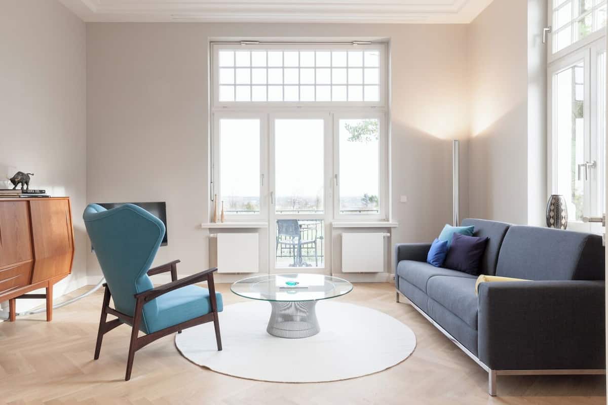 A modern living room with a round glass coffee table, a blue armchair, a grey sofa, and a wooden cabinet.