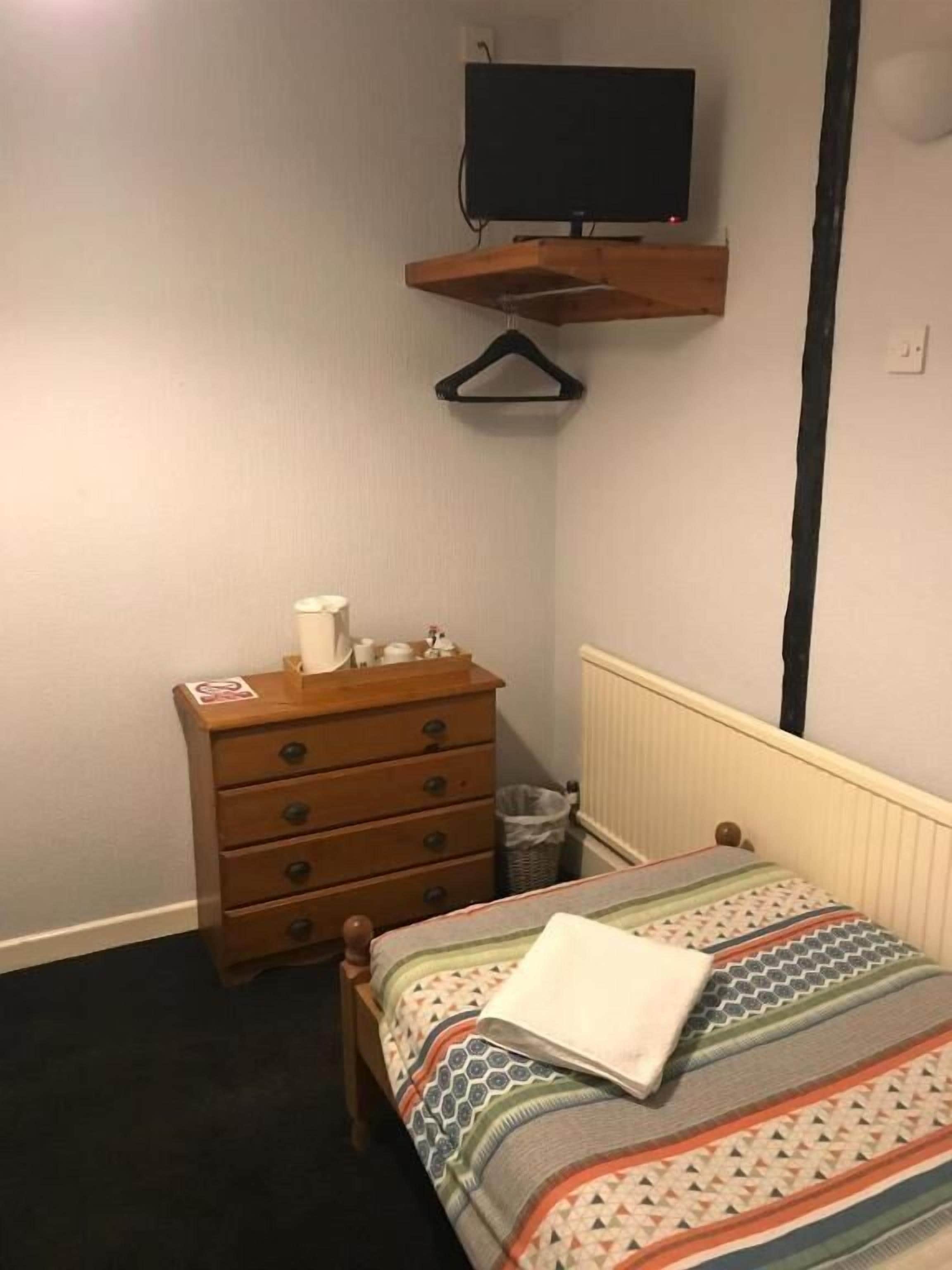 A bedroom with a wooden dresser, a hanging TV, a radiator, and a patterned bedspread.