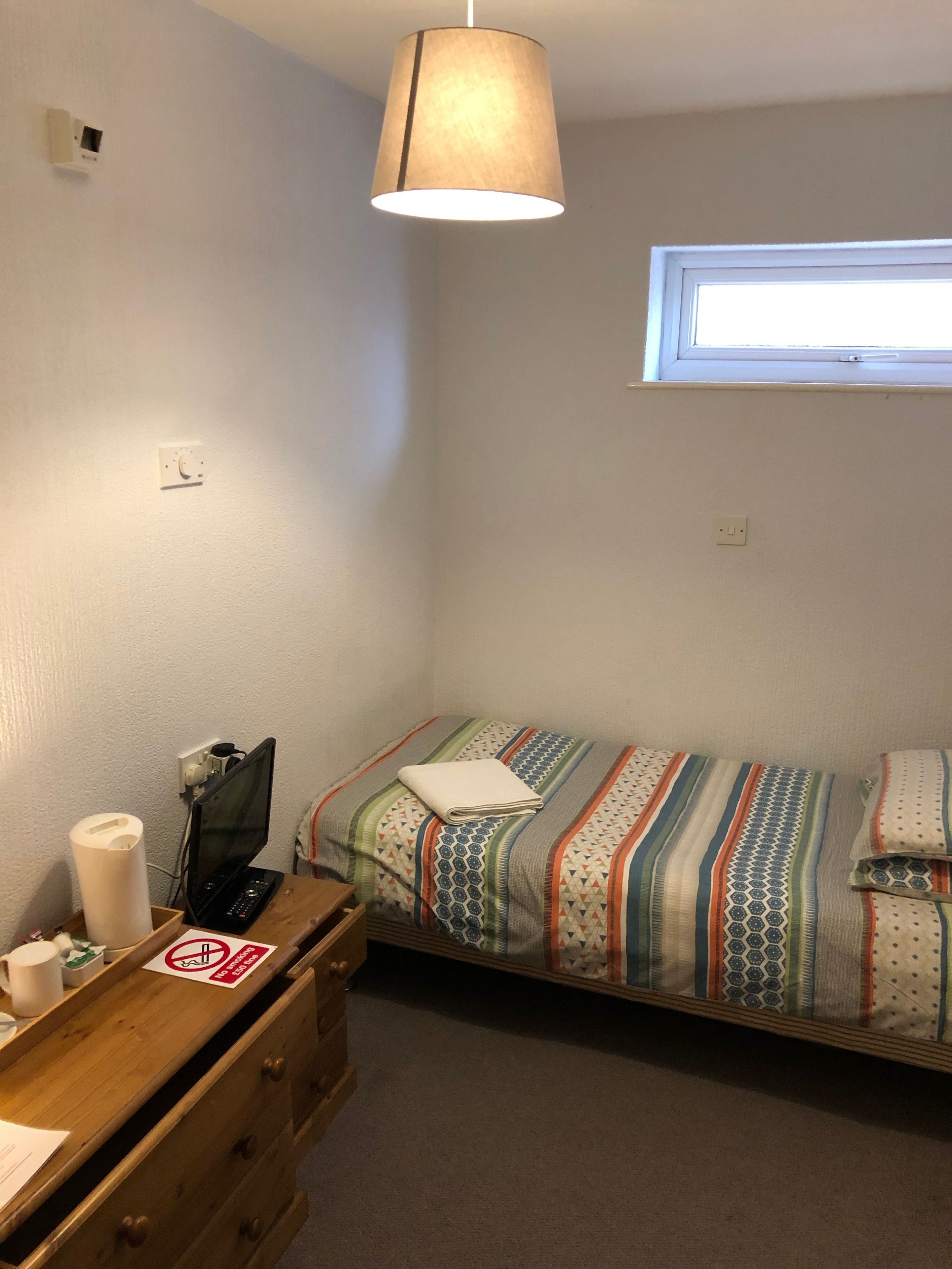 A small, single-bed room with a window, a desk with a computer, and a lamp.