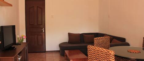 Una sala de estar con un sofá marrón oscuro, una mesa de centro de madera, una silla de mimbre, un televisor sobre un mueble de madera y una puerta con manija.
