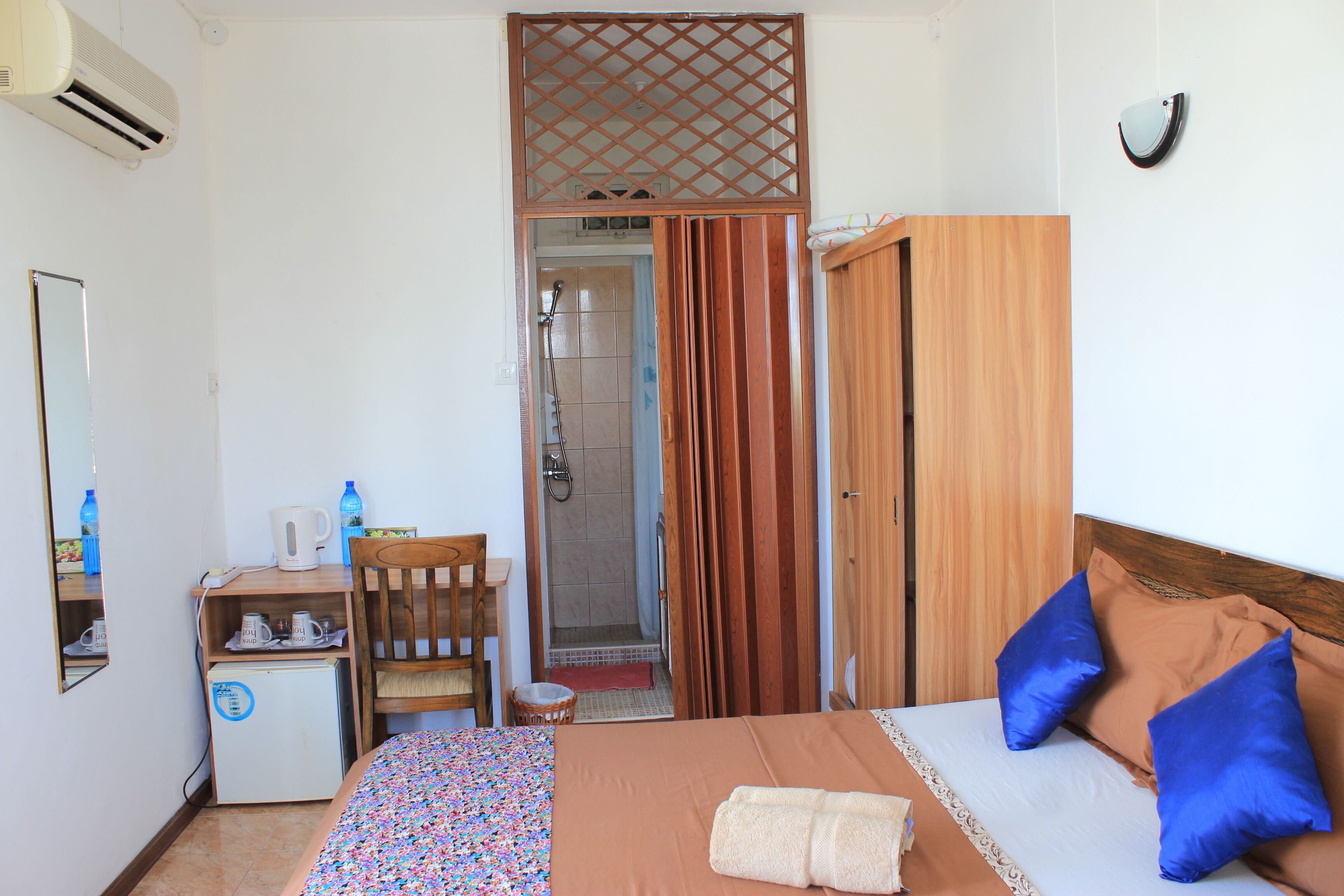 A hotel room with a bed, a desk with a refrigerator, a chair, a shower area, and a wardrobe.