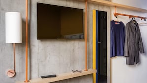 A modern room with a mounted TV, a shelf with a remote, and a clothing rack with garments. - ibis budget Lyon Est Beynost (Beynost)