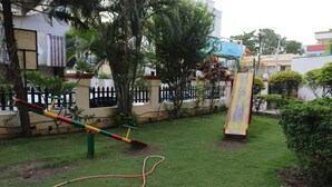A playground with a slide and swings, surrounded by a low stone wall and greenery. - Hotel Riviera Executive (Chhatrapati Sambhajinagar)