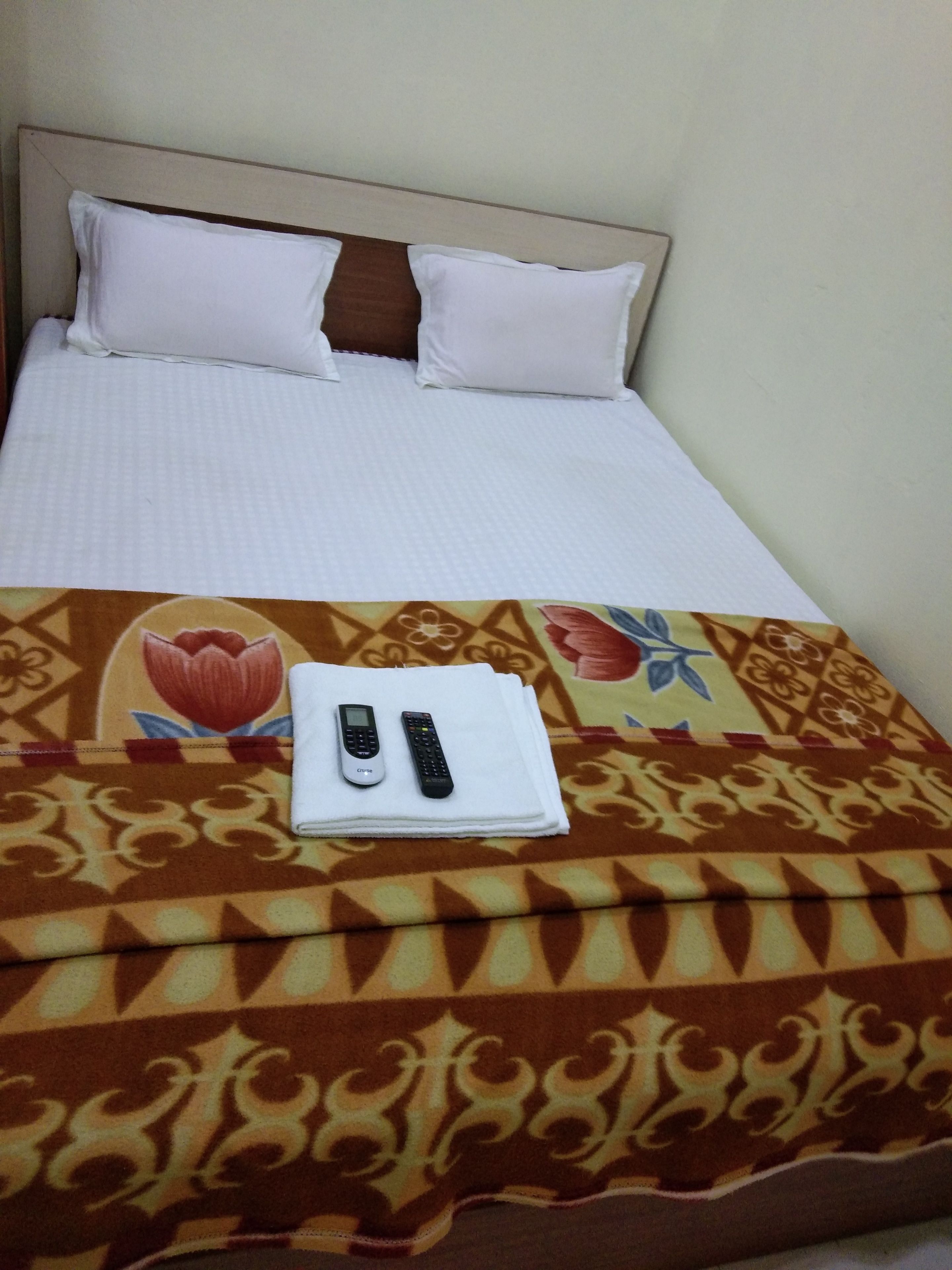 Una cama bien hecha con sábanas blancas y dos almohadas, una manta estampada y dos controles remotos sobre una toalla doblada.