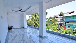 Ein geräumiges Zimmer mit Marmorboden, Deckenventilator und einem Balkon mit Metallgitter, der auf ein Wohngebiet mit Häusern und Palmen blickt.