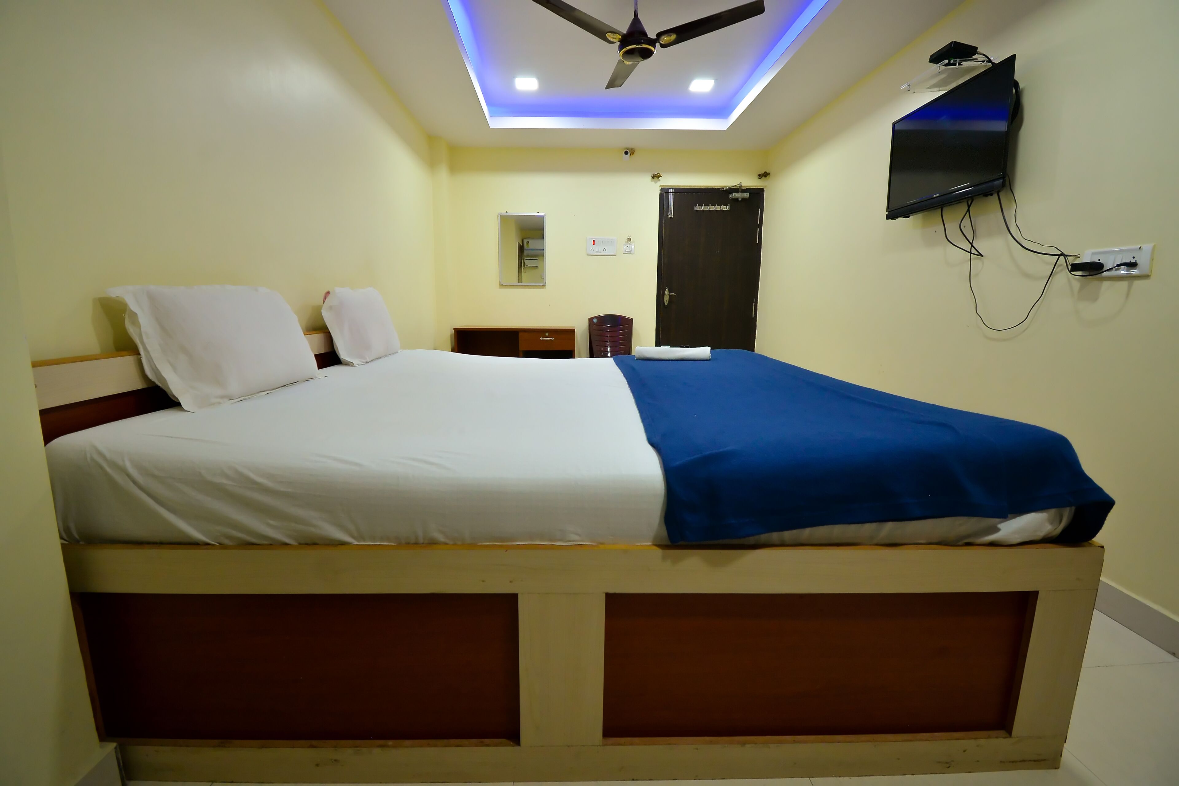 A bed with a blue and white bedspread, a ceiling fan, a mounted TV, and a door with a lock.