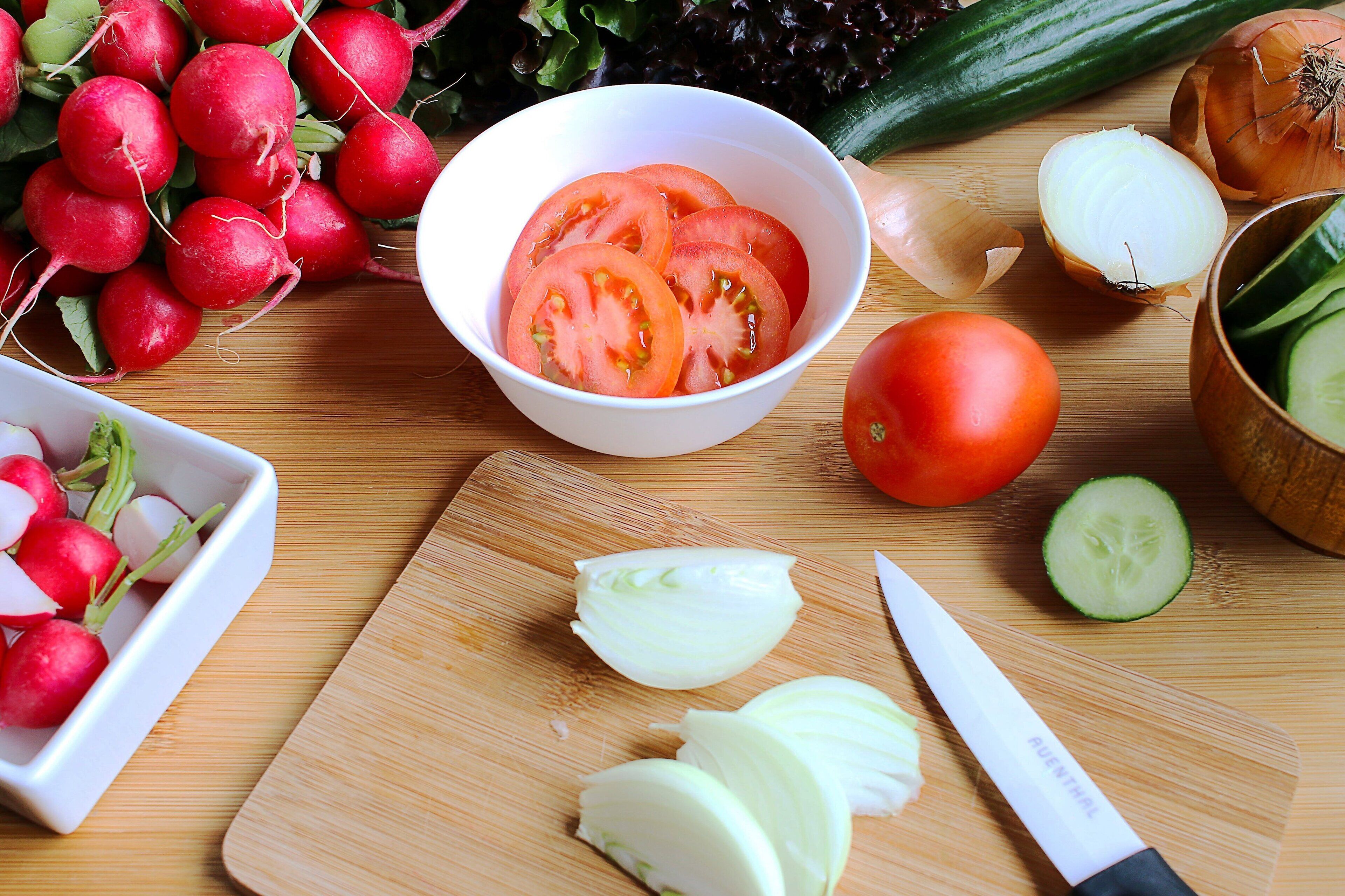 A wooden cutting board with sliced vegetables, a knife, and a bowl of sliced tomatoes.