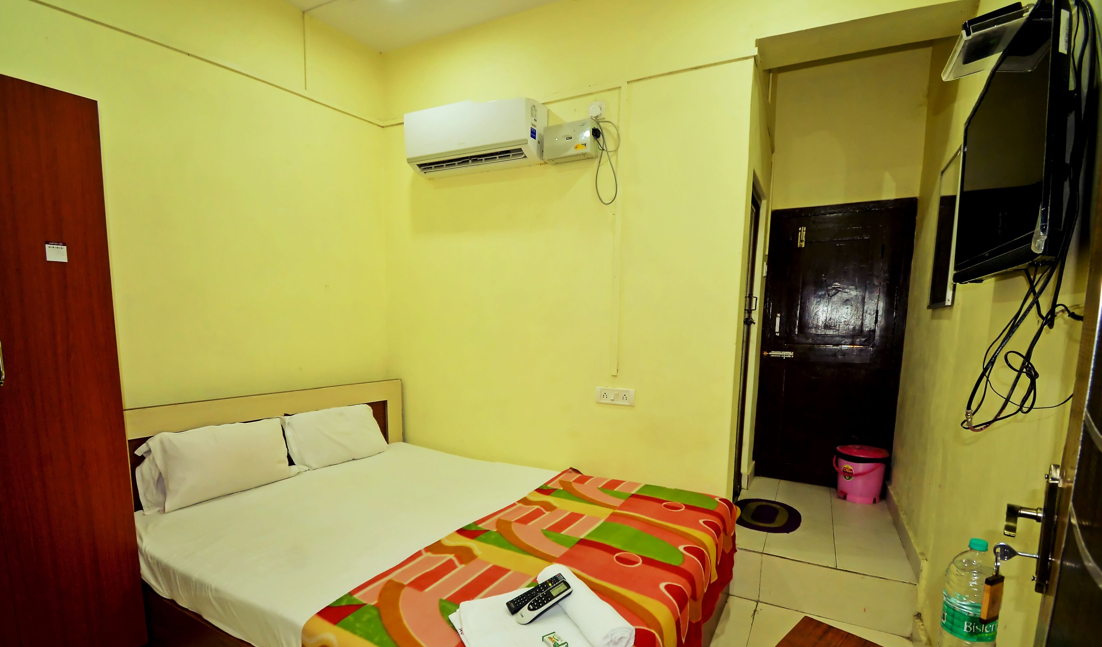 A hotel room with a bed, an air conditioner, a television mounted on the wall, and a bucket on the floor.