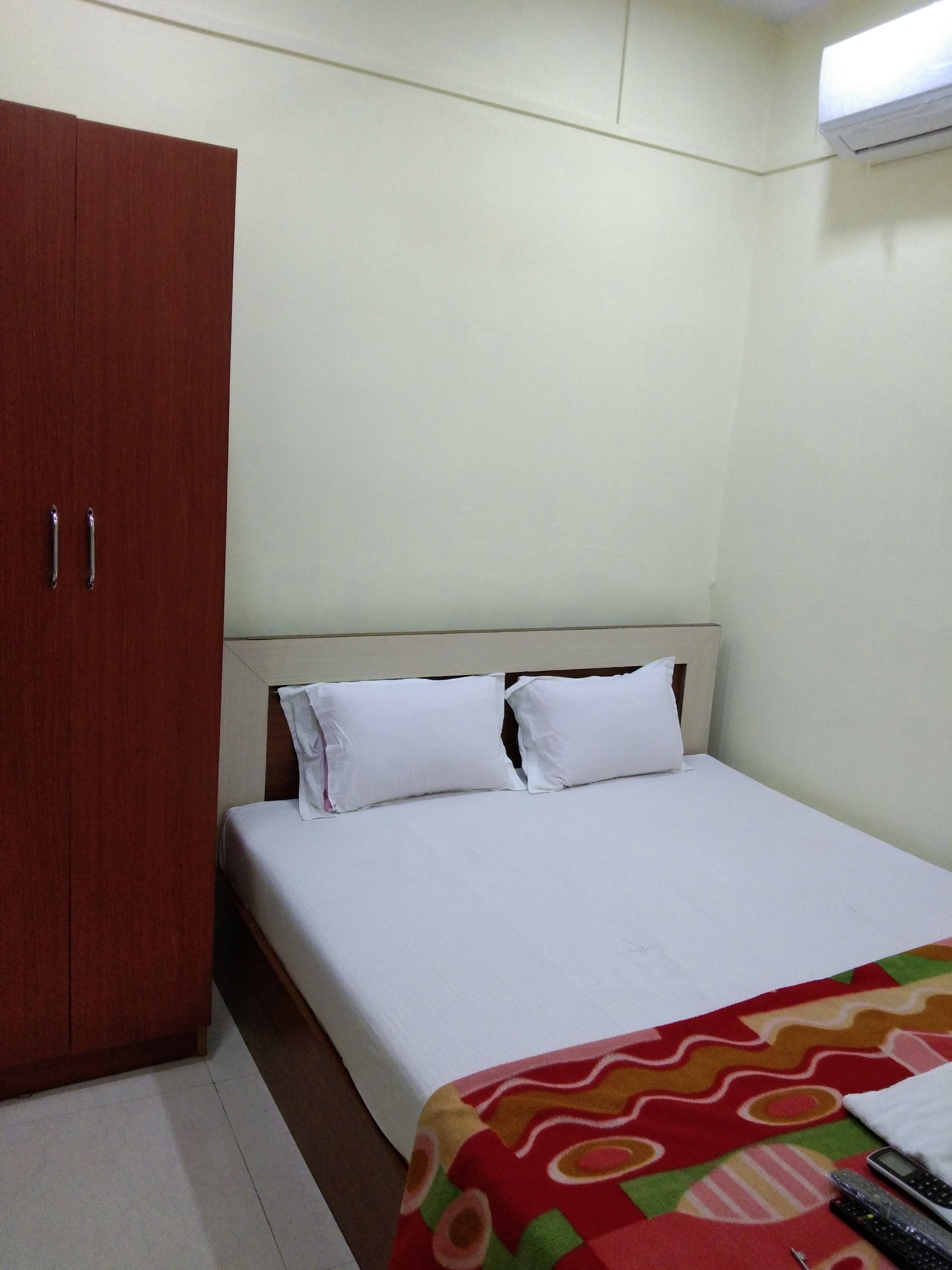 A single-bed room with a wooden wardrobe, a bed with white linens, and a colorful blanket.