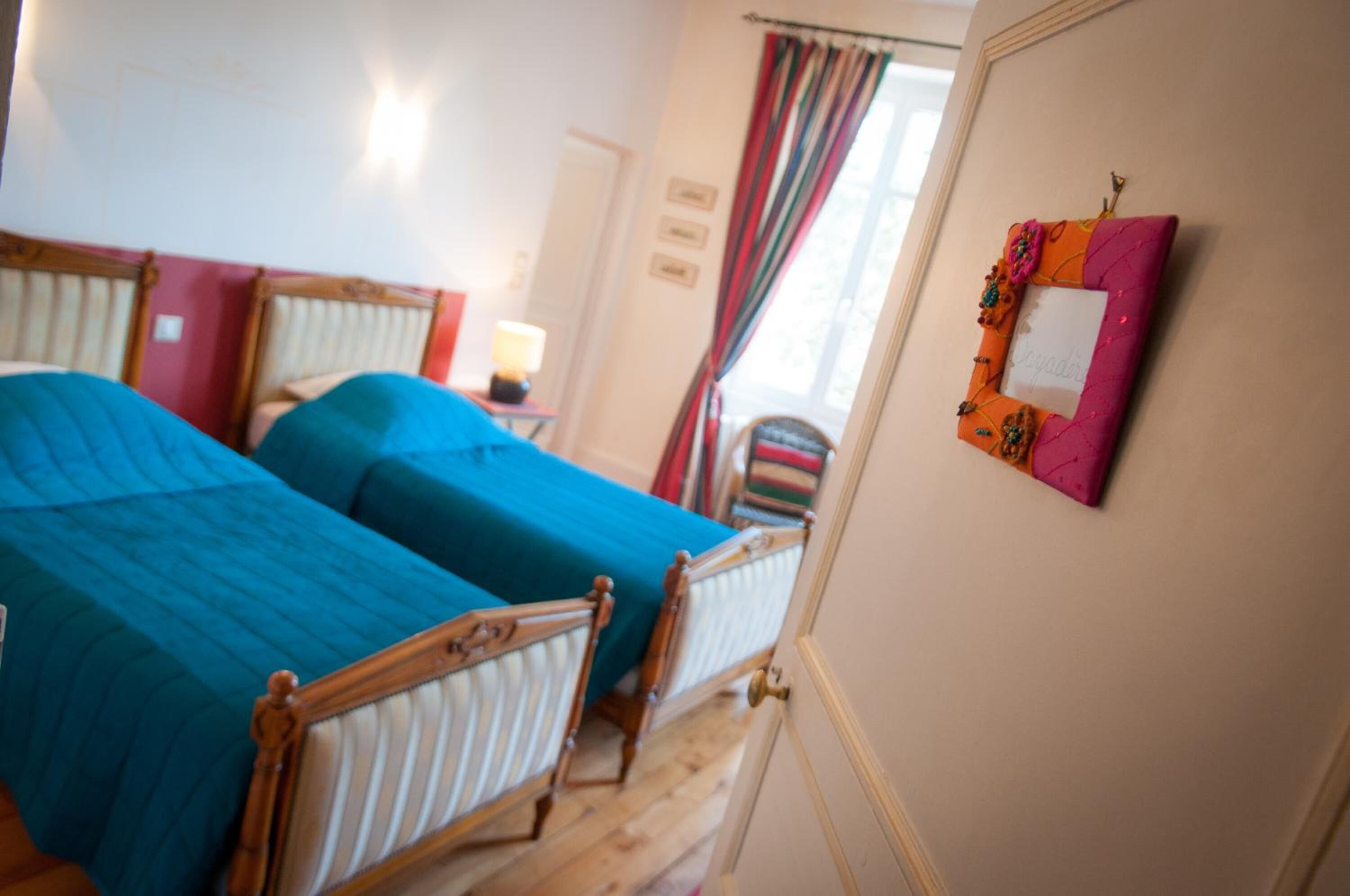 A room with two beds, a small table with a lamp, and a colorful wall decoration.