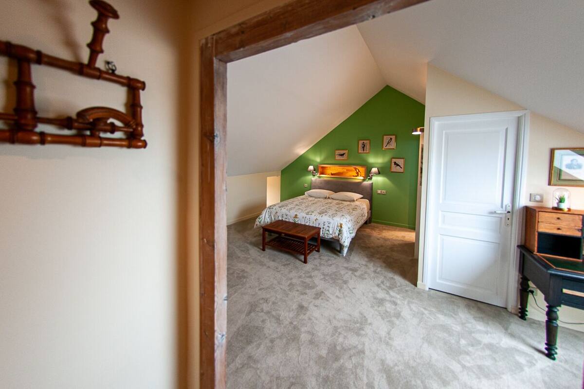 A bedroom with a sloped ceiling, a bed with a patterned bedspread, a wooden bench, and a desk with a lamp.