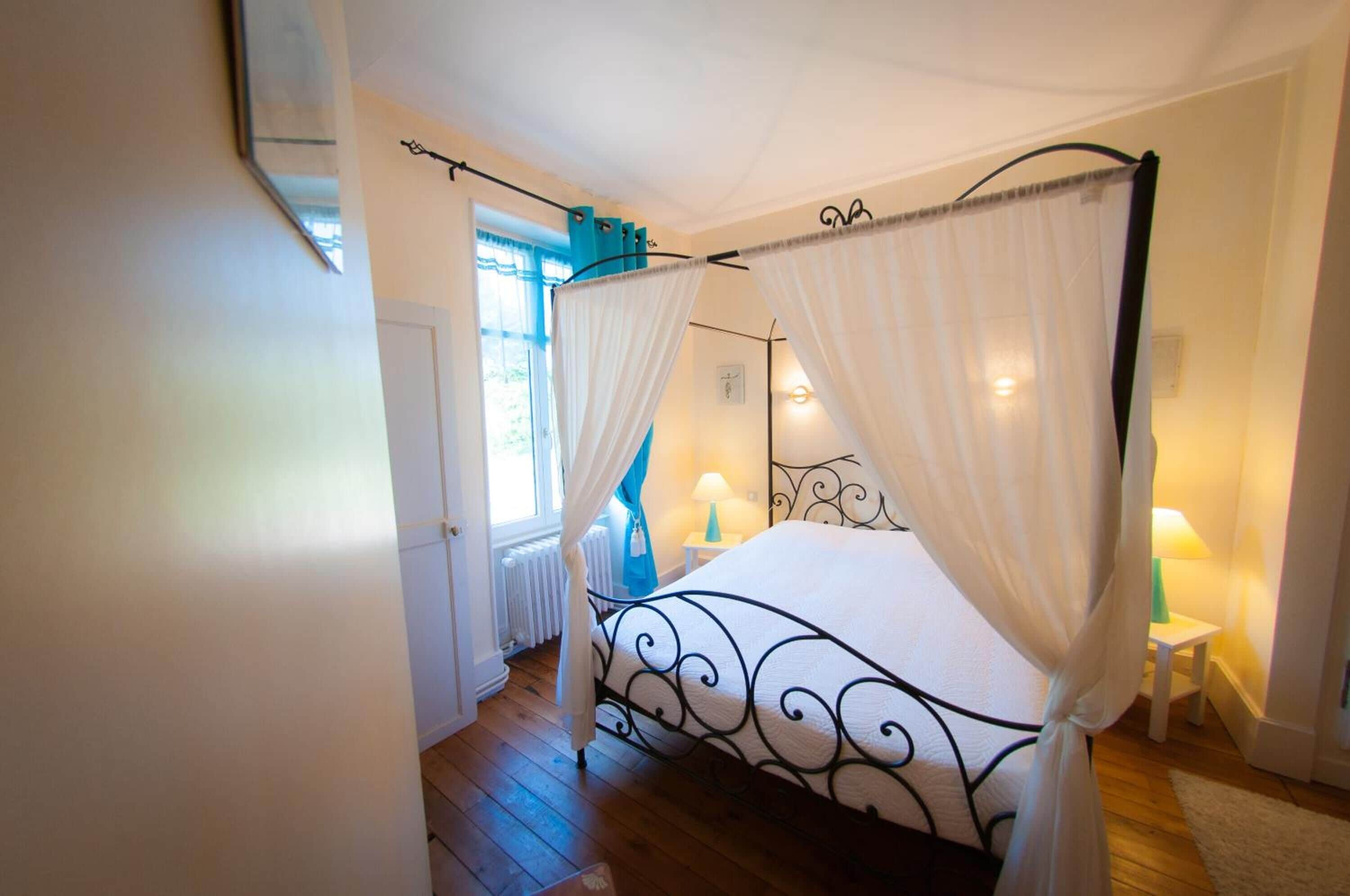 A bedroom with a four-poster bed, white curtains, a small bedside table with a lamp, and a window with blue curtains.