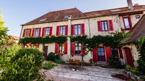 A two-story building with red shutters, a tiled roof, and a small garden area with a table and chairs. - Au Puy des Vérités (Lapalisse)