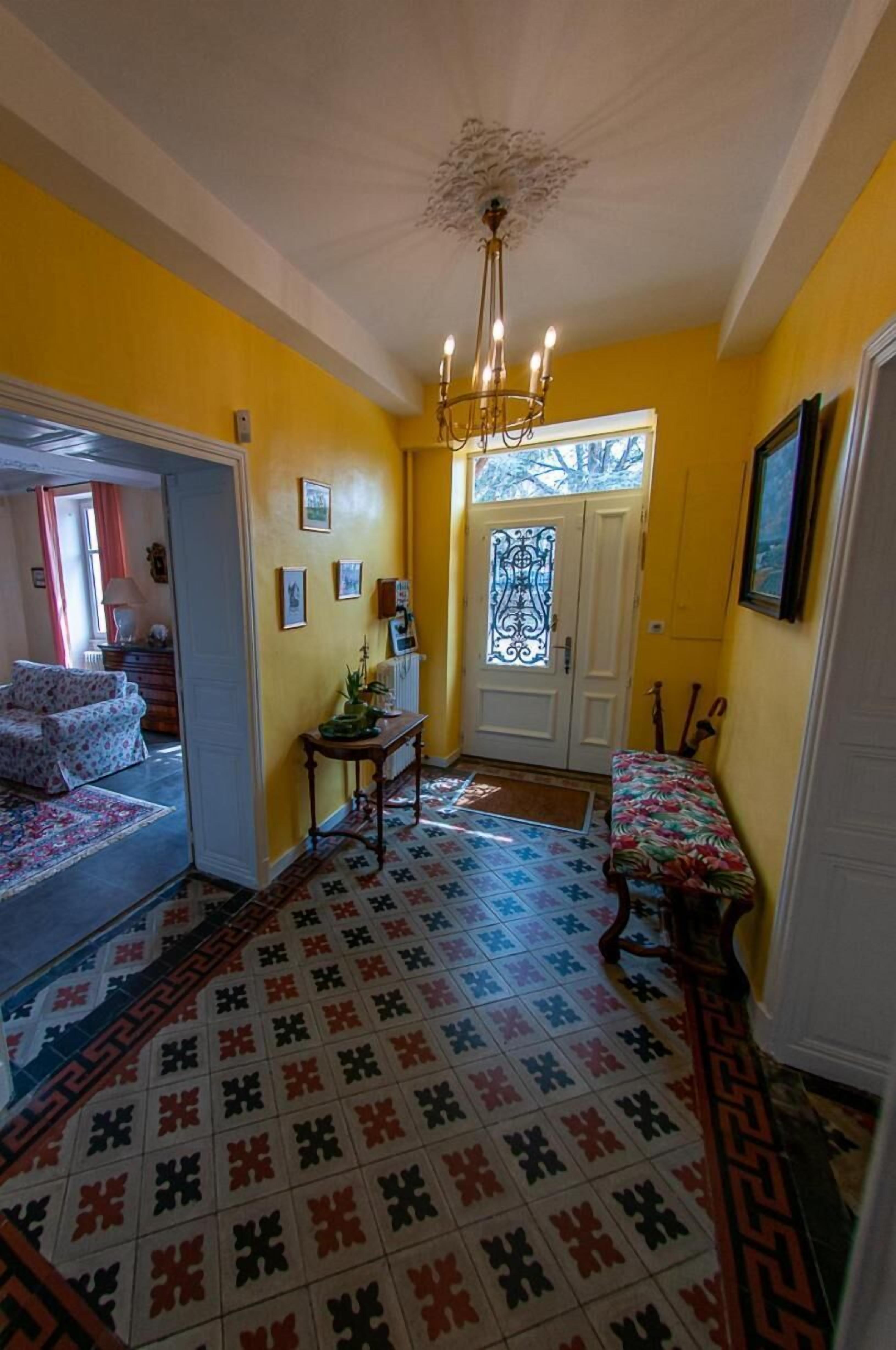 A hallway with patterned floor tiles, a bench, a door with a decorative glass panel, and a chandelier.