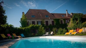 A swimming pool with lounge chairs and a large building with a sloped roof in the background. - Au Puy des Vérités (Lapalisse)