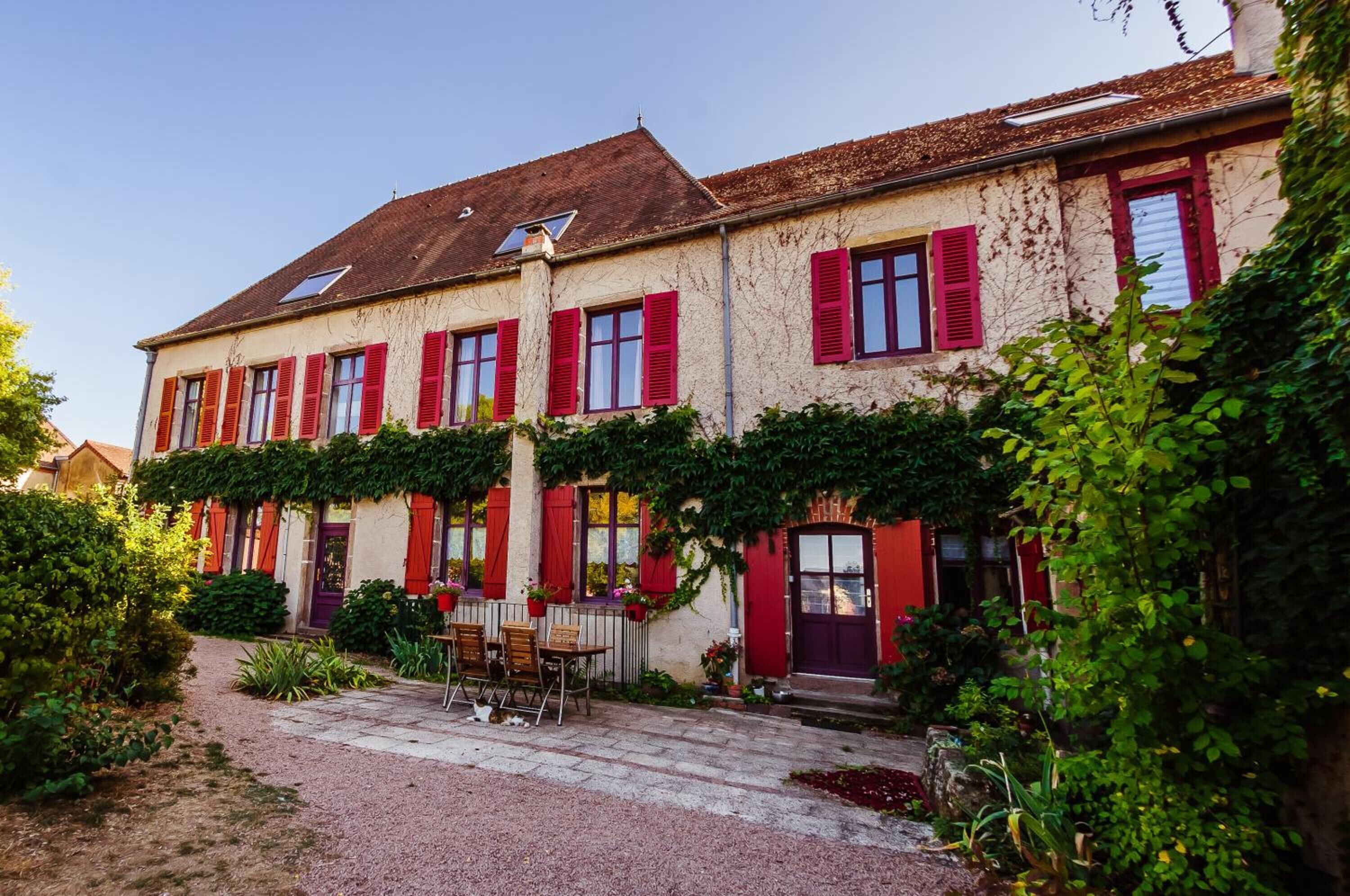 A two-story building with red shutters, a gravel pathway, and a small outdoor seating area with tables and chairs.