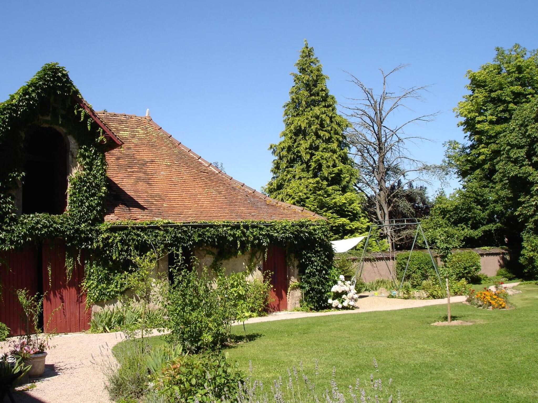 Un bâtiment de style rustique, doté d’un toit en tuiles, entouré d’un jardin bien entretenu où poussent différentes espèces de plantes et où s’étend une pelouse.