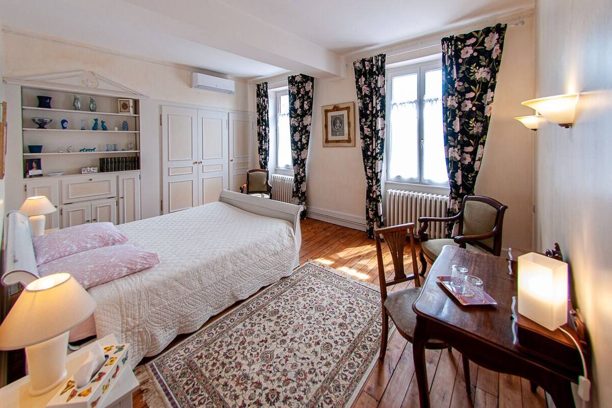 A bedroom with a bed, bedside table, lamp, and a chair. There is a window with curtains, a radiator, and a shelf with decorative items.