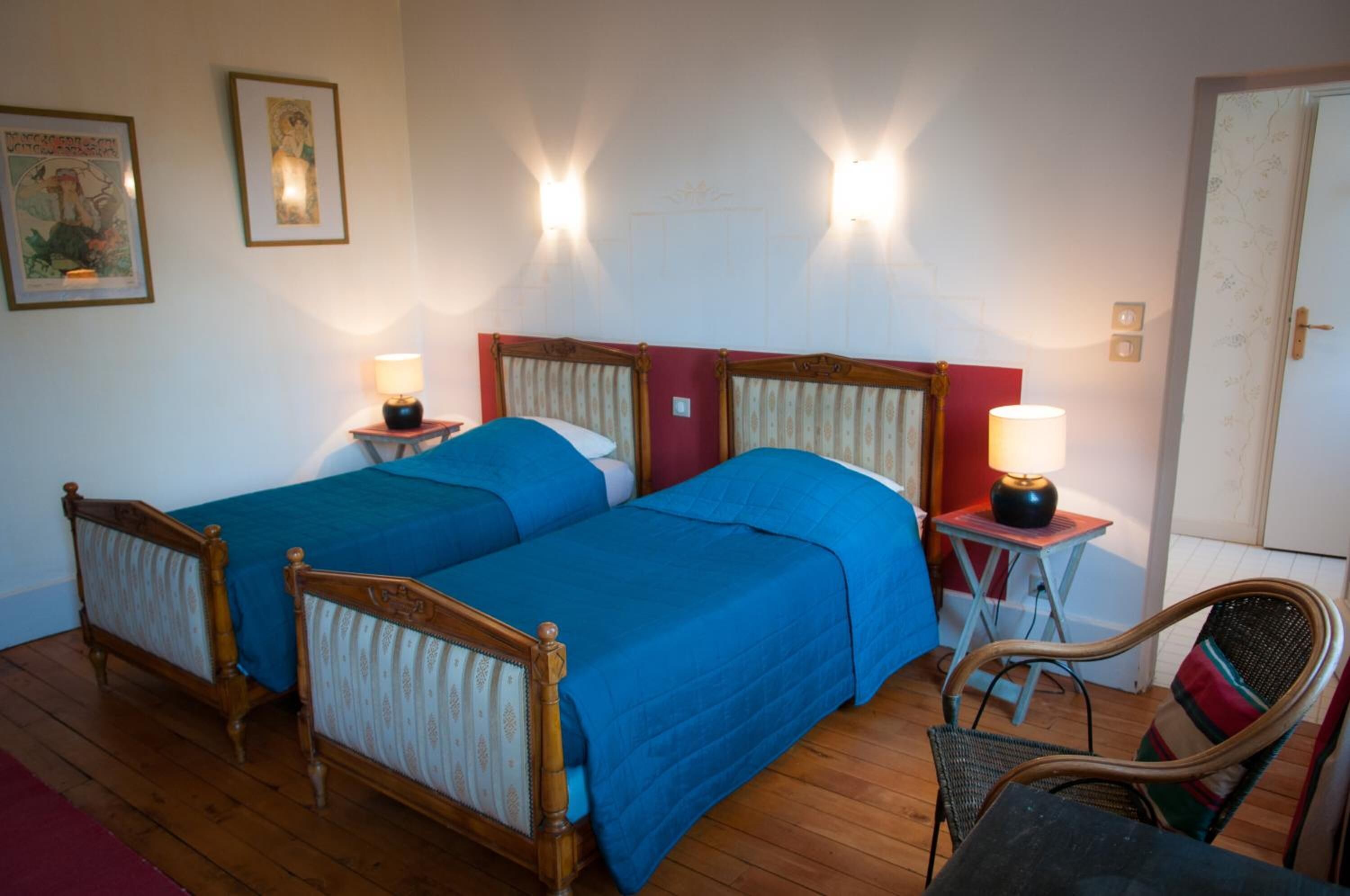 Een tweepersoonsbed met houten frame en blauw beddengoed, twee nachtkastjes met lampen, een rieten stoel en een deur met bloemenprint.