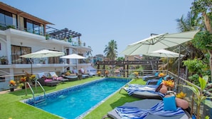 A pool area with lounge chairs and umbrellas, surrounded by greenery and a building with multiple balconies. - Nitya Resort (Calangute)