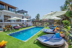 A pool area with lounge chairs and umbrellas, surrounded by greenery and a building with multiple balconies.