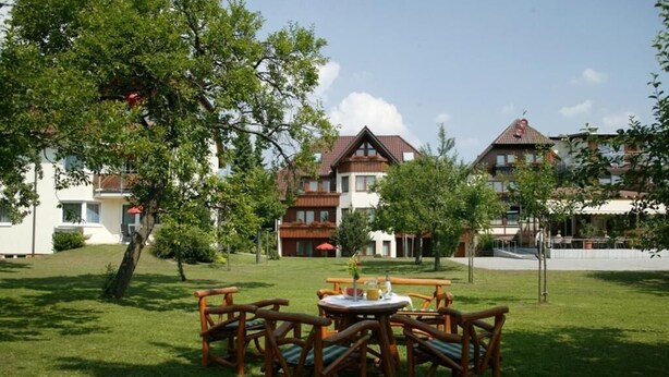 Une cour avec une table et des chaises en bois, entourée d’une végétation luxuriante et d’arbres.
