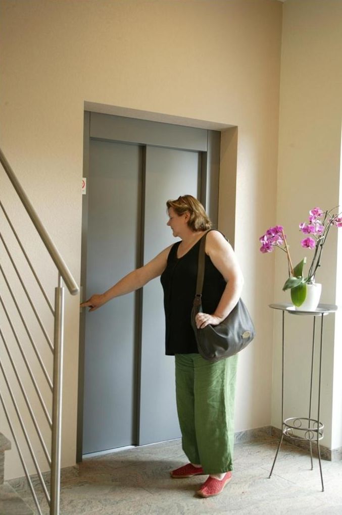 A woman standing at an elevator door in a hallway with a potted plant nearby.