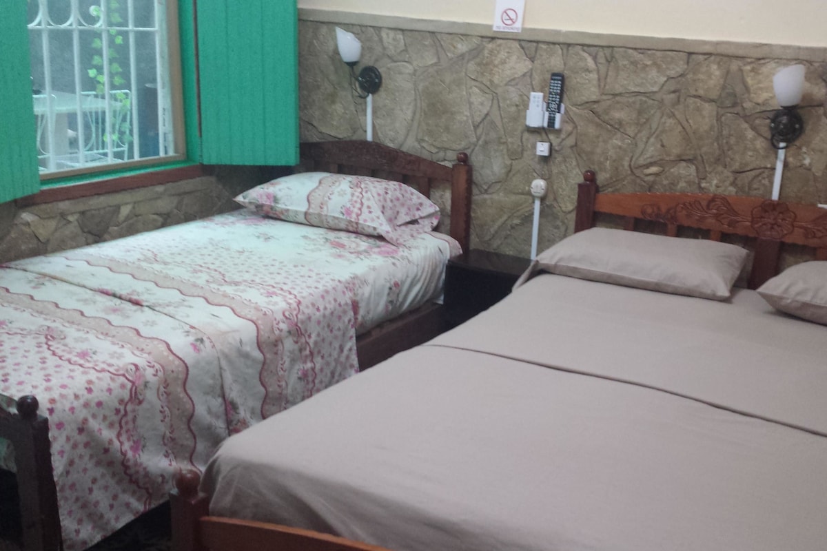 A room with two single beds and one double bed, a stone wall, a window with bars, and a wall-mounted light fixture.