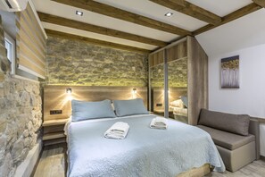 Quarto com parede de pedra, vigas de madeira, cama com colcha e um banco.