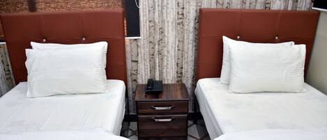 Una habitación de hotel con dos camas, cada una con ropa blanca y cabeceros marrones. Entre las camas hay una mesita de noche de madera con un teléfono y un cajón.