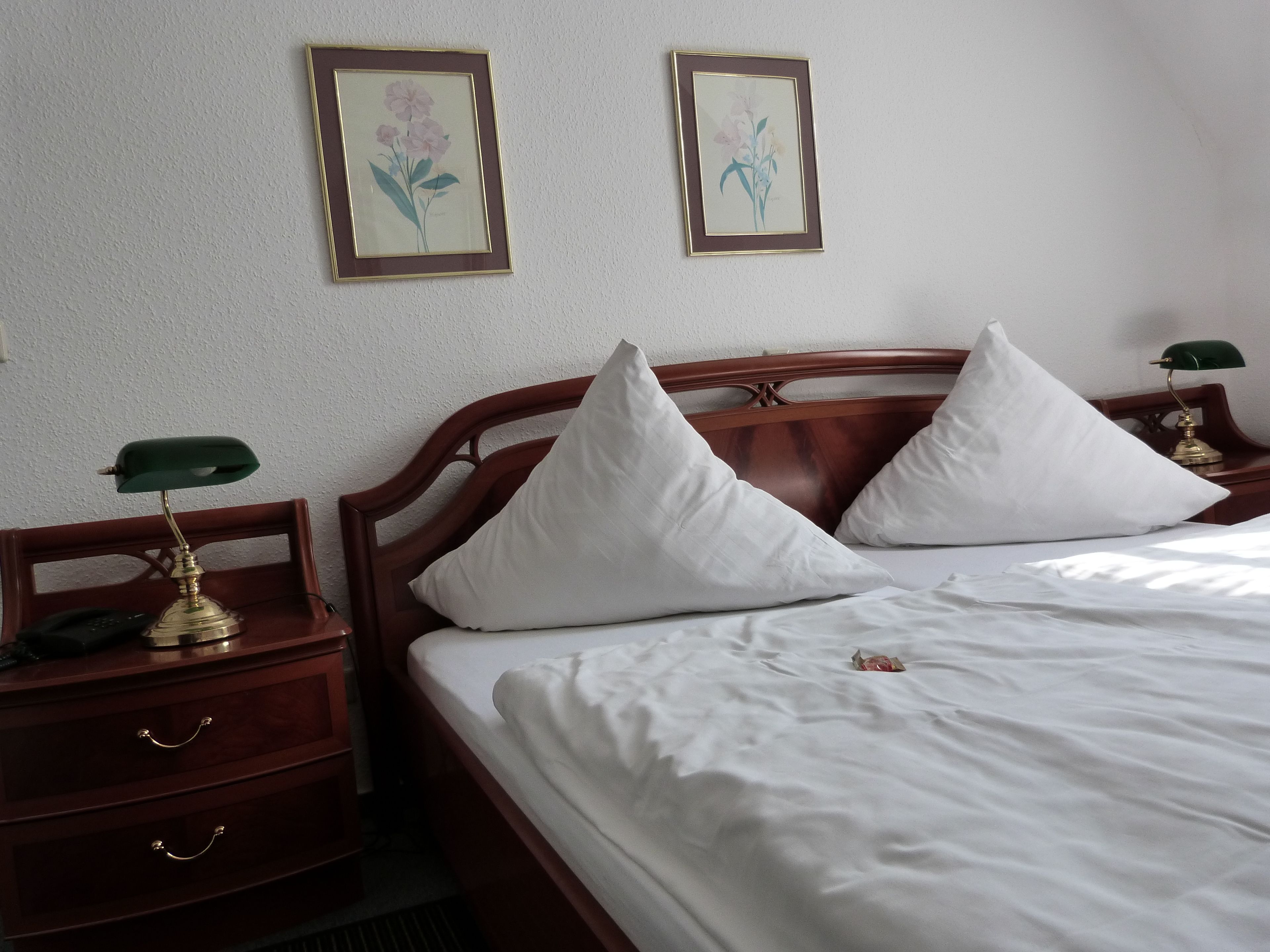 A neatly made bed with white linens and two pillows, a wooden headboard, a bedside table with a lamp, and two framed floral pictures on the wall.