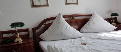 Una cama bien hecha con sábanas blancas y dos almohadas, cabecera de madera, una mesita de noche con una lámpara, y dos cuadros enmarcados con motivos florales en la pared.