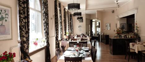 Un comedor con una larga mesa dispuesta para una comida, flanqueado por ventanas altas con cortinas, una araña y una zona de cocina al fondo.