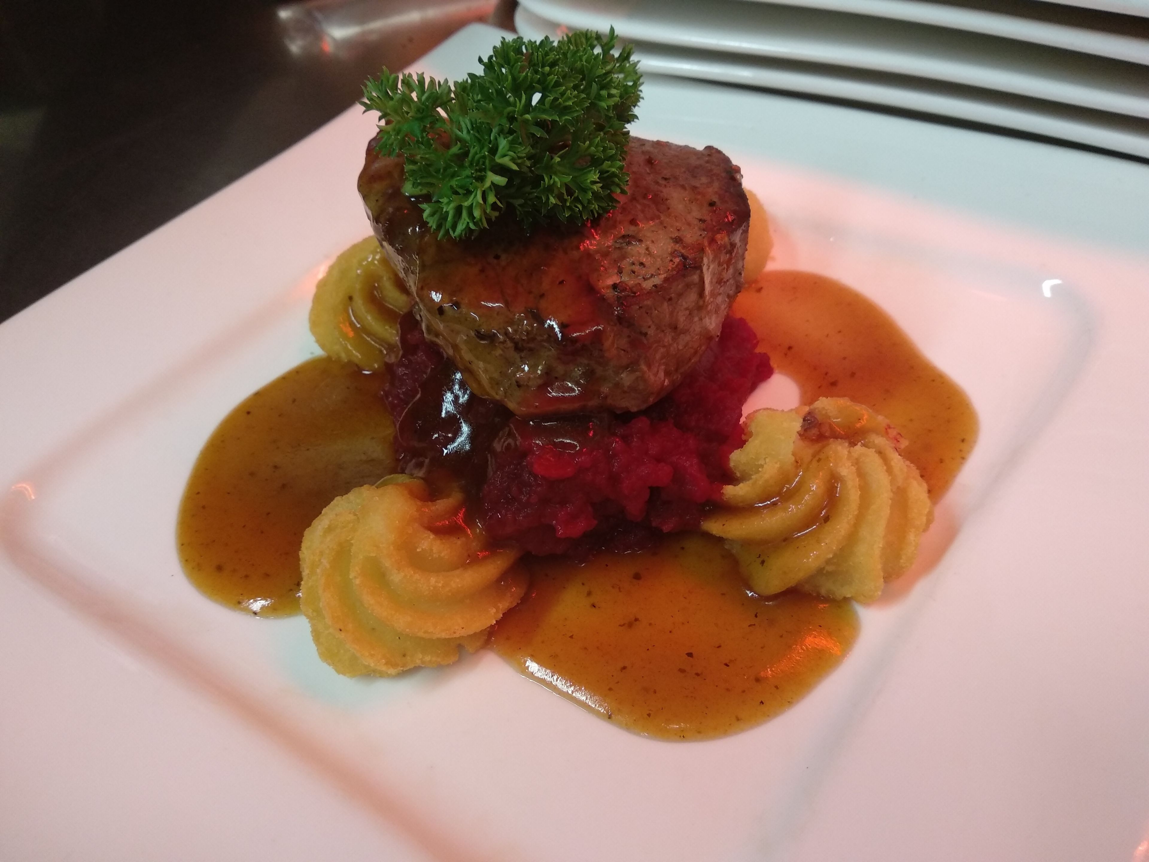 Un plato con un guiso de carne como pieza central, adornado con perejil, rodeado de un puré rojo y una salsa marrón.