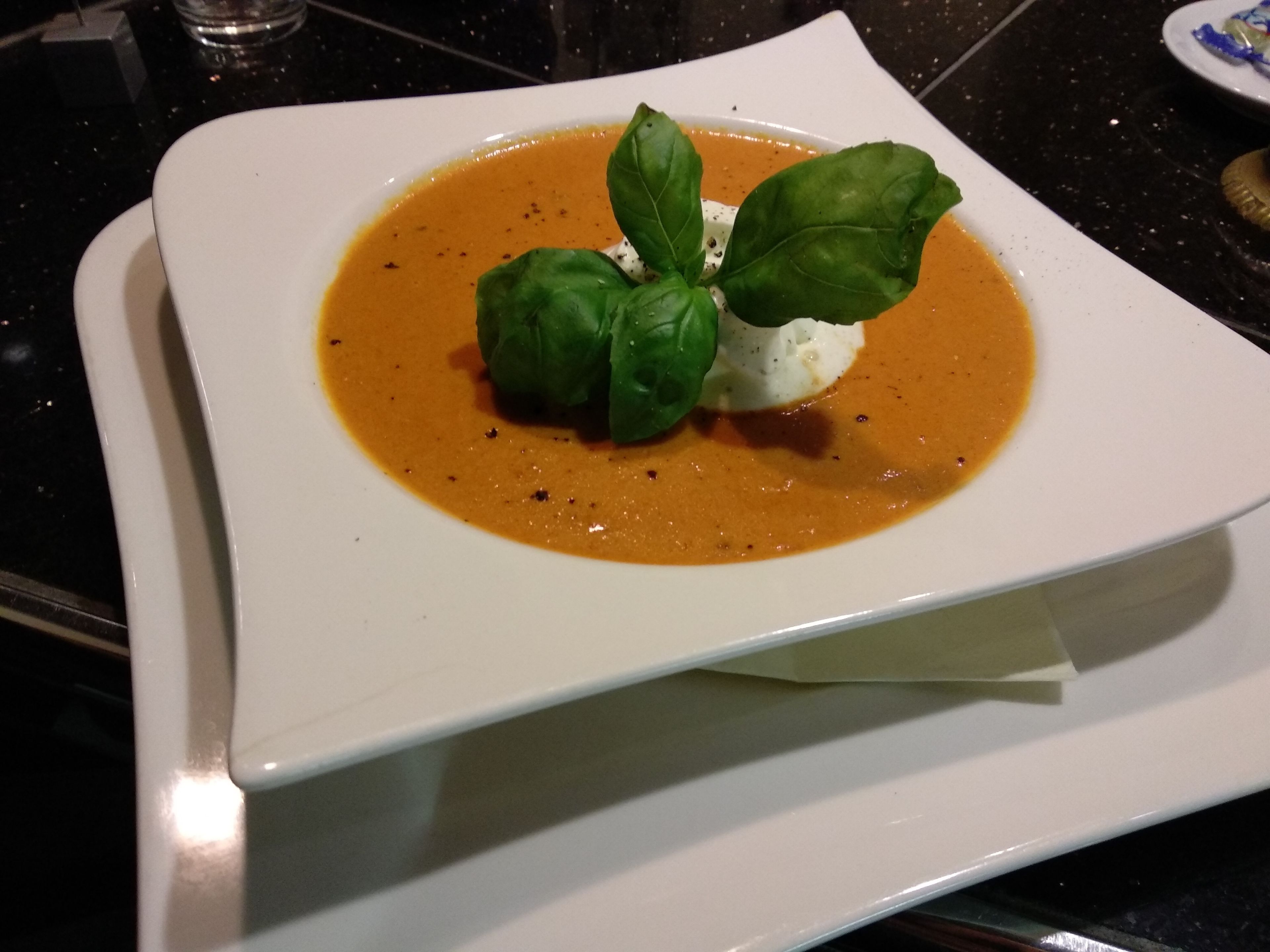 A square white plate with a creamy orange soup, two green leaves, and a dollop of white cream.