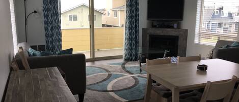 A living room with a wooden table and chairs, a sofa, a glass coffee table, a fireplace, and a TV mounted on the wall.