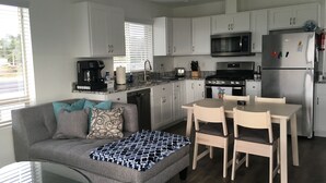 Een moderne keuken met witte kasten, een roestvrijstalen koelkast, een magnetron en een fornuis. Er is een eettafel met stoelen en een bank met een decoratieve plaid.