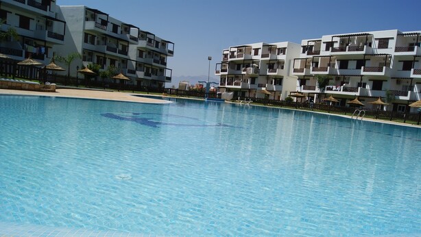 A swimming pool with tiled edges, surrounded by multi-story buildings with balconies and outdoor seating areas. - Résidence Mirador Golf Cabo Negro (M'diq)