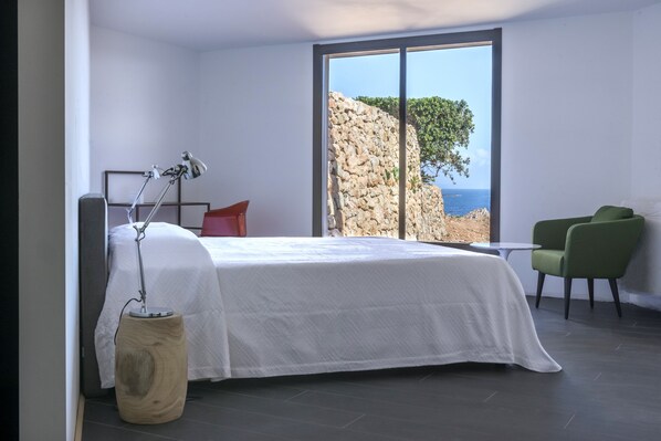 A modern bedroom with a large bed, a desk, a chair, and a view of the sea through a large window.