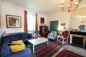 Uma sala de estar com um sofá azul, uma mesa de centro de madeira, um tapete vermelho estampado, uma poltrona azul e uma lareira.